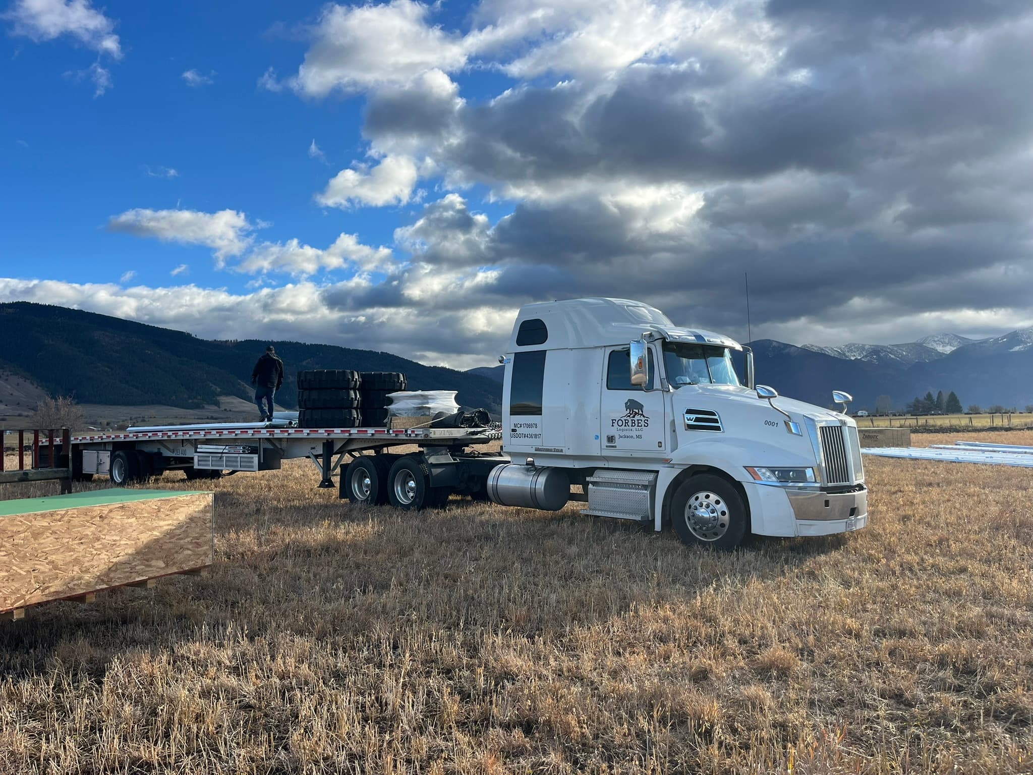 Forbes Logistix flatbed truck loading freight on a Western plain with Rocky Mountains in the background